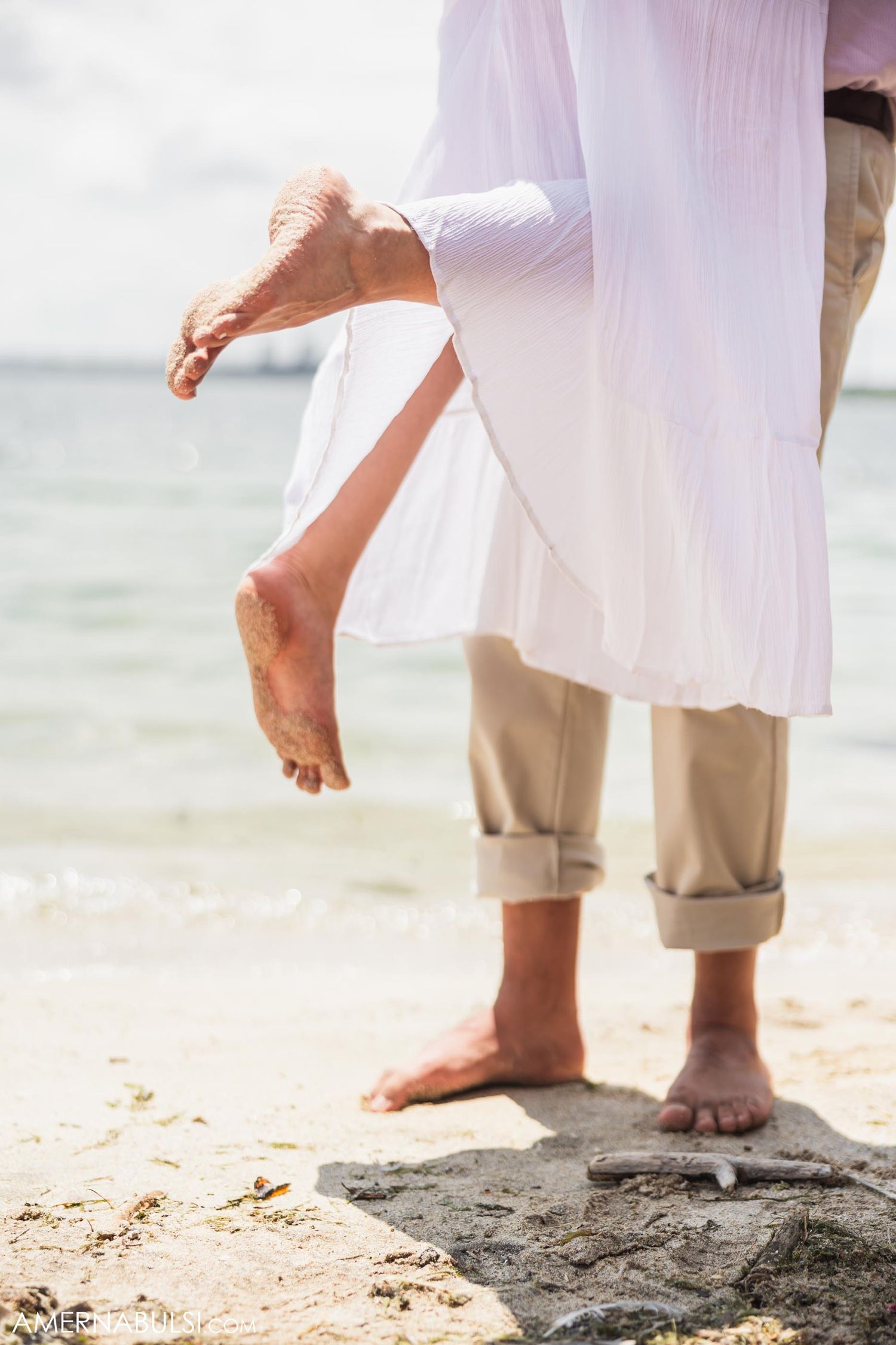 Bare feet of couple standing on beach rocks with flowing white dress fabric.