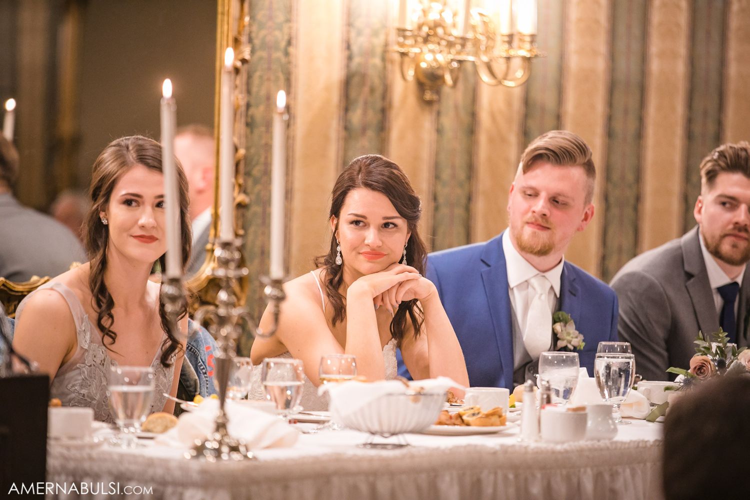 Wedding reception table setting with guests seated in elegant dining room with chandelier lighting.