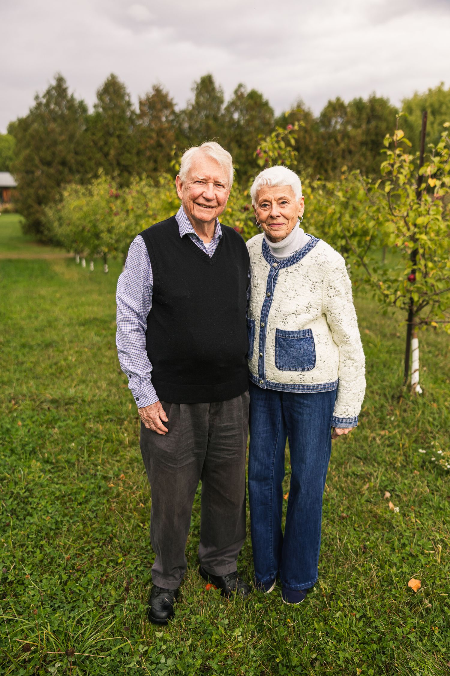 Elderly couple standing together in an apple orchard on an overcast autumn day.