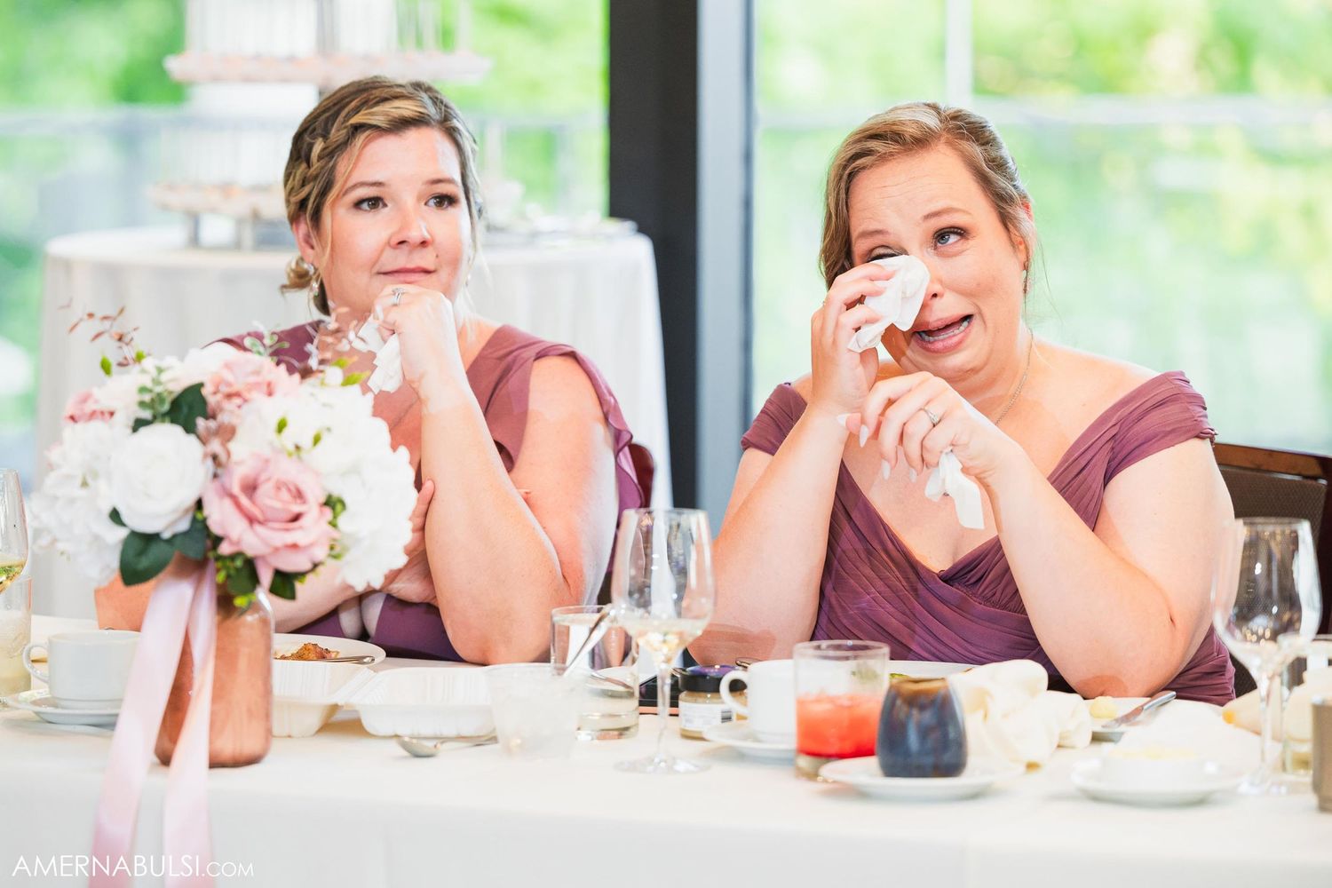 Two guests at a wedding reception share an emotional moment while wiping away tears.