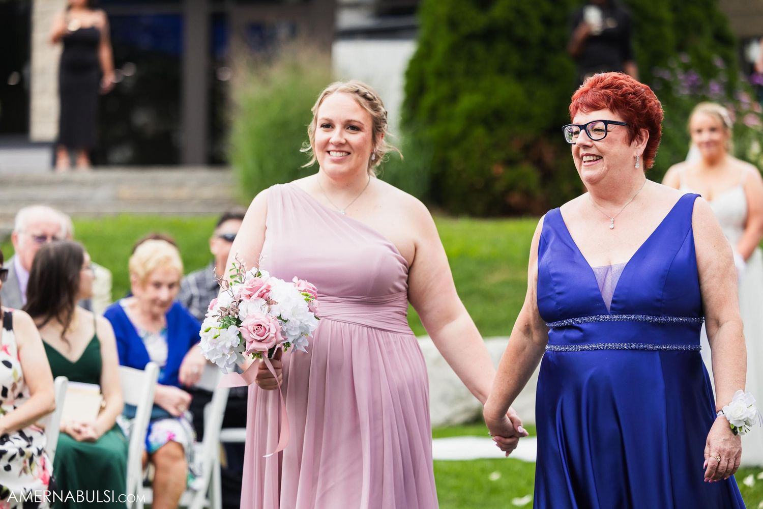 Two bridesmaids in mauve and royal blue dresses walk together holding bouquets during an outdoor wedding ceremony.