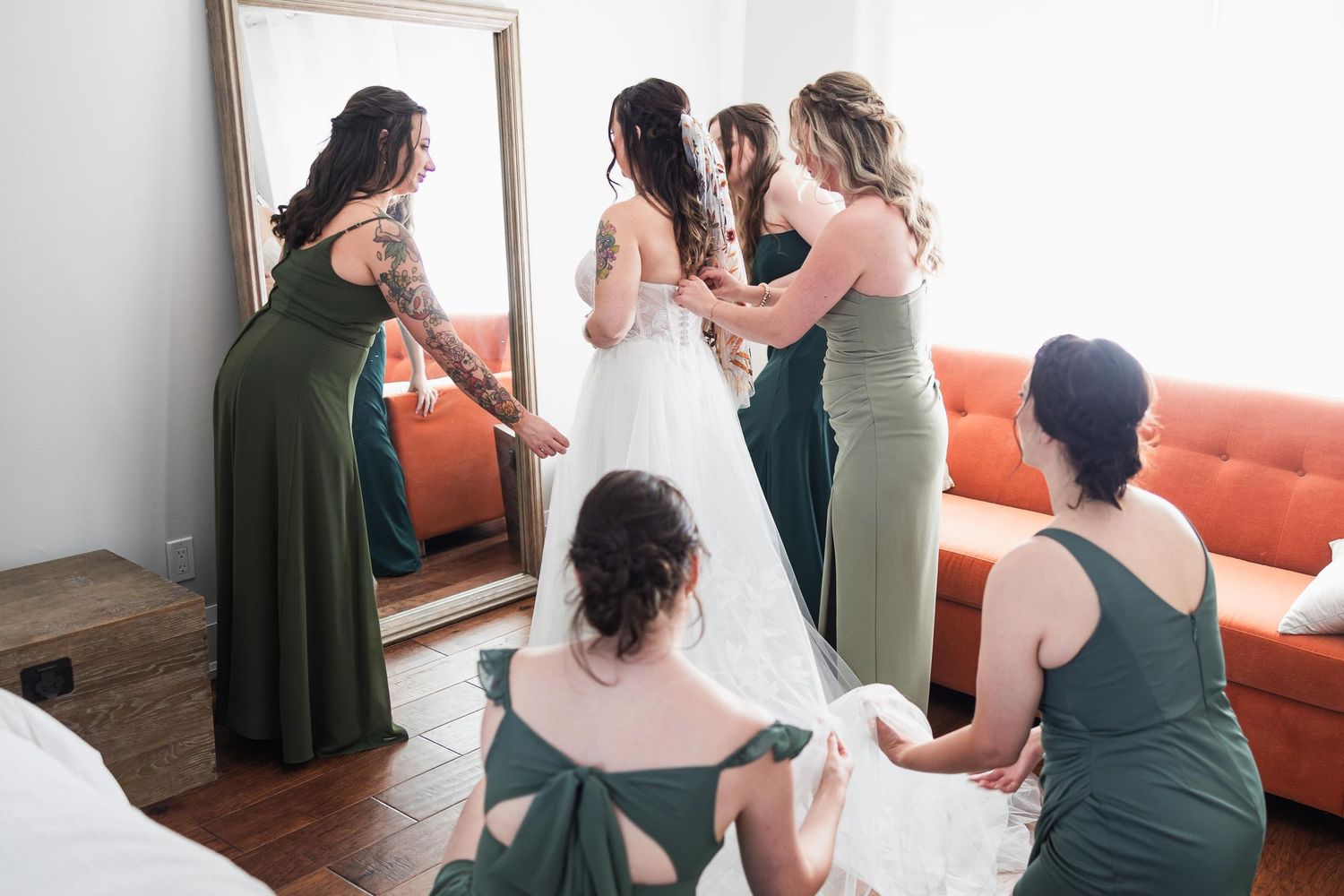 Bridesmaids in green dresses help with wedding preparations while gathered around a mirror and couch.
