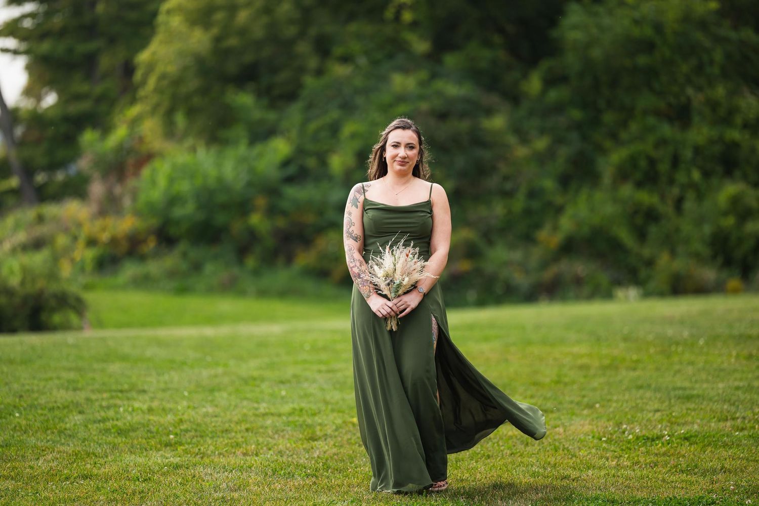Bridesmaid poses in flowing sage green dress on lawn with bouquet.