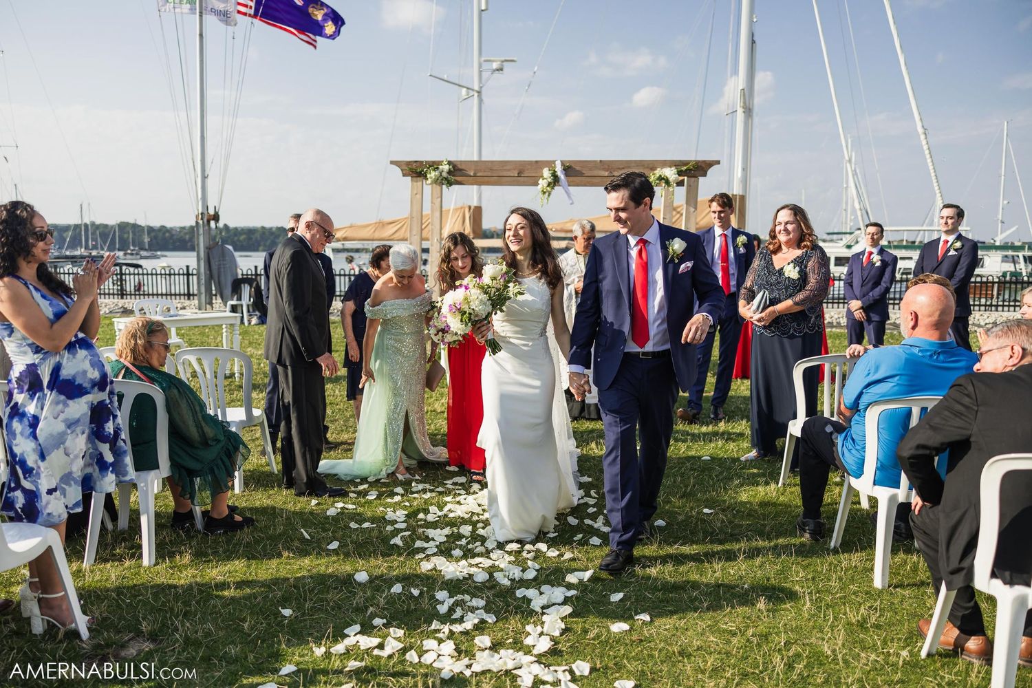 Outdoor wedding ceremony taking place on a grassy lawn near a marina with guests seated and flower petals scattered.