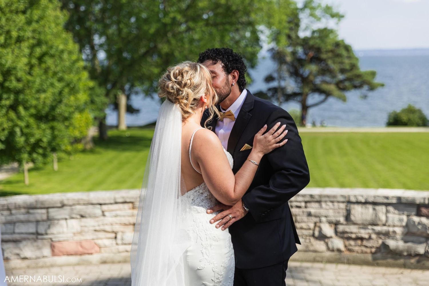 A romantic wedding kiss between newlyweds on a stone terrace overlooking a scenic waterfront.