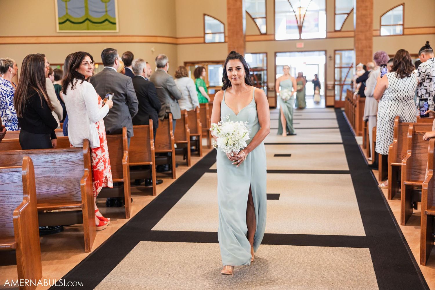An attendant in a pale blue dress proceeds down the church aisle holding a floral bouquet.