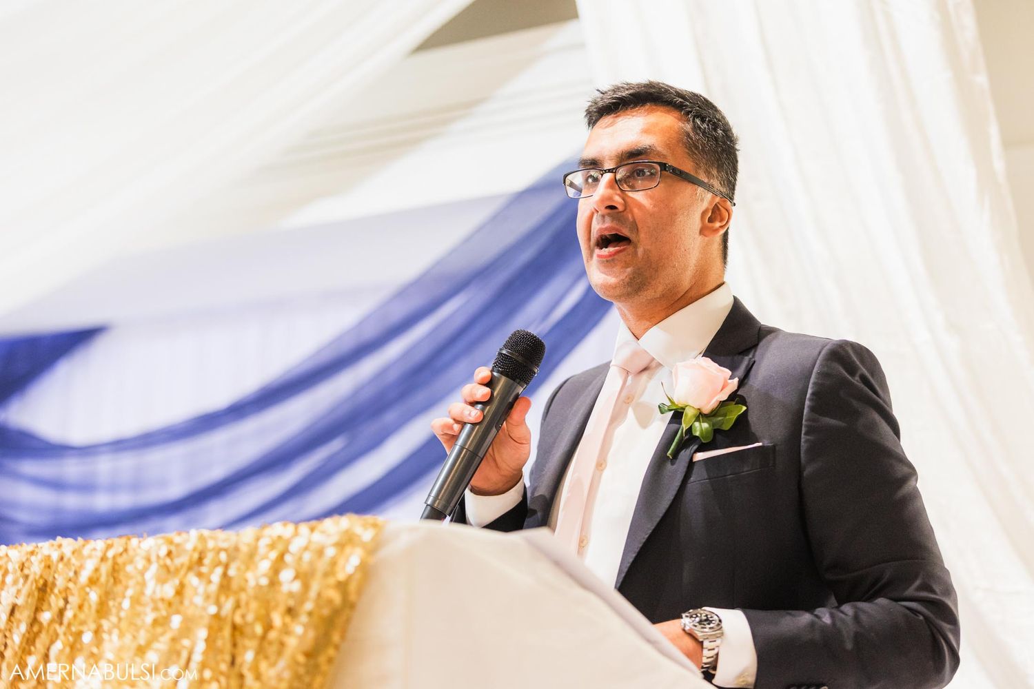 Wedding speaker delivers speech at reception podium with blue draping and gold sequin backdrop.