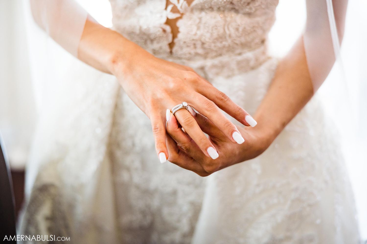 Close-up of bride's hands showing off wedding ring against white lace dress.