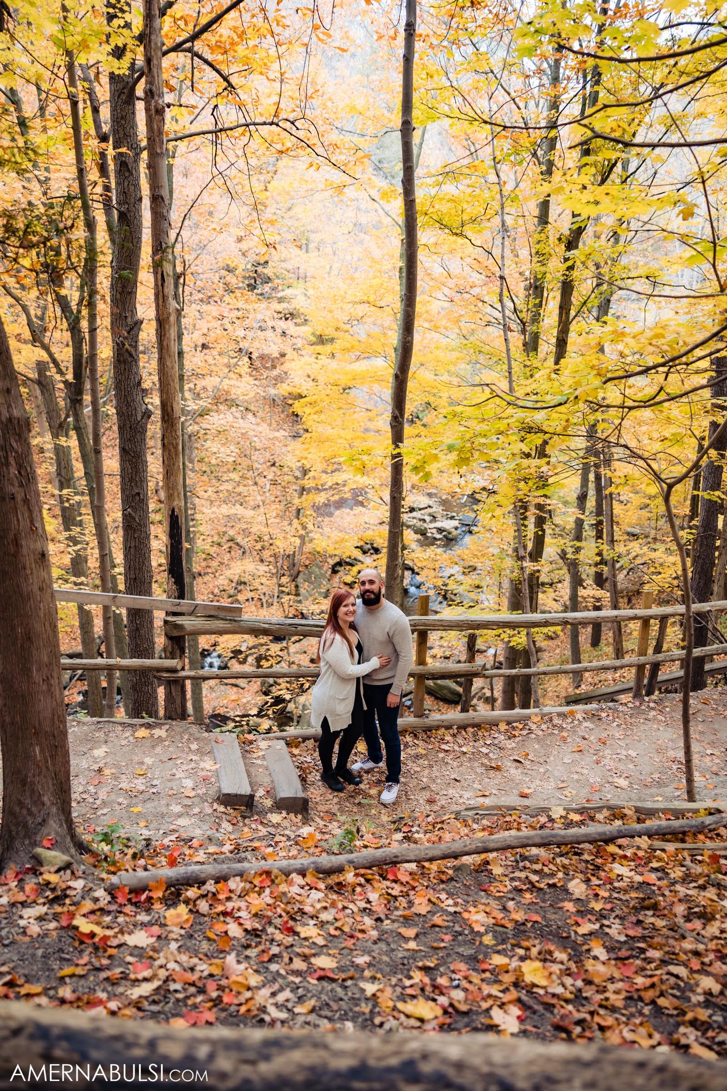 Couple stands together on wooden walkway surrounded by yellow autumn trees.