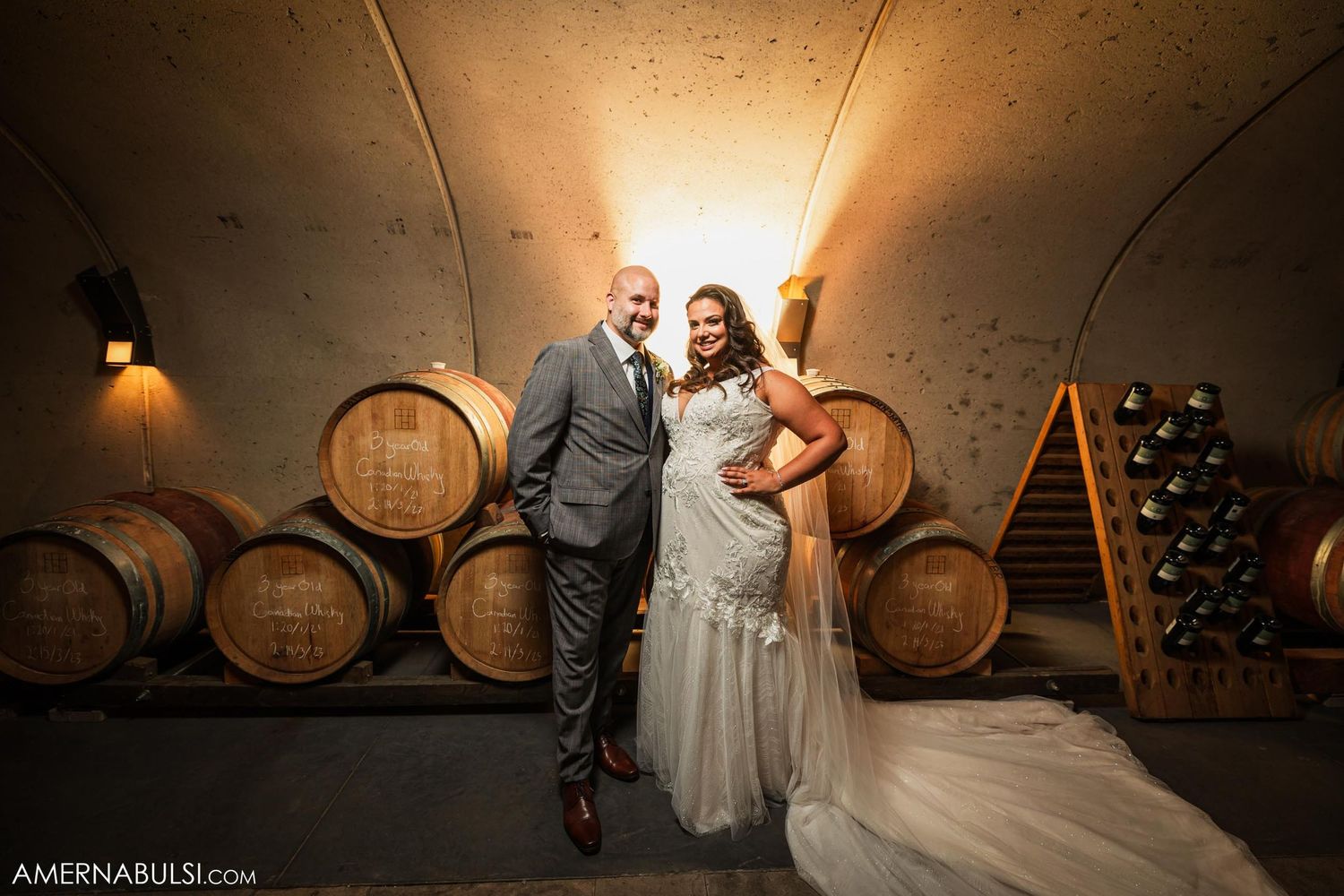 Elegant couple poses in rustic wine cellar with oak barrels.