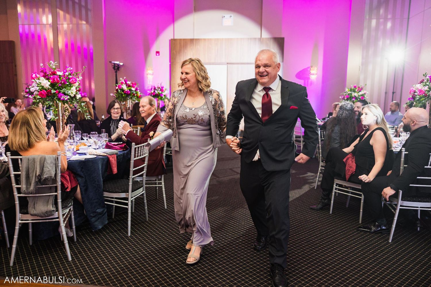 Wedding guests joyfully dancing between reception tables with pink mood lighting.