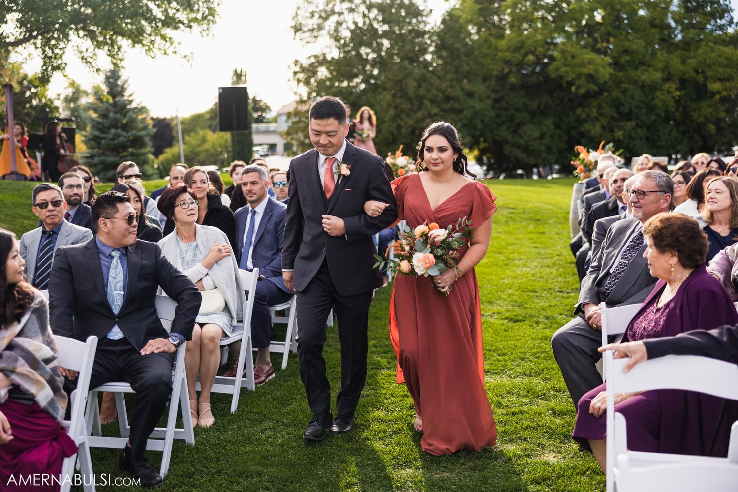 A bridesmaid in mauve walks down an outdoor aisle escorted by a groomsman.