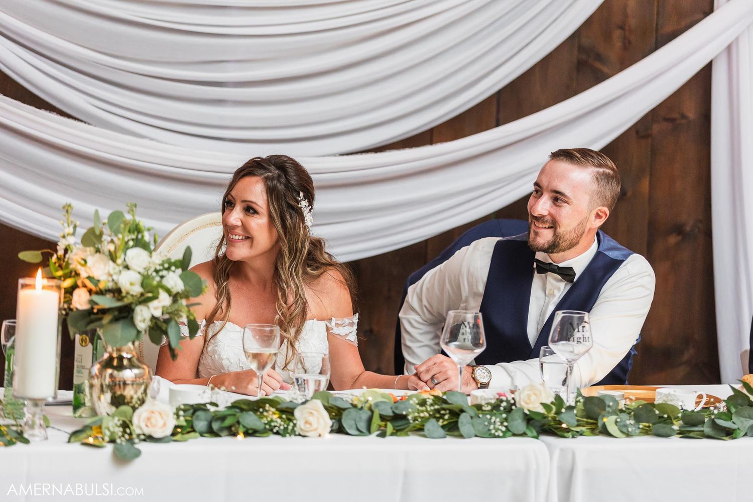 A couple sits at their decorated head table with white draping and greenery garland.