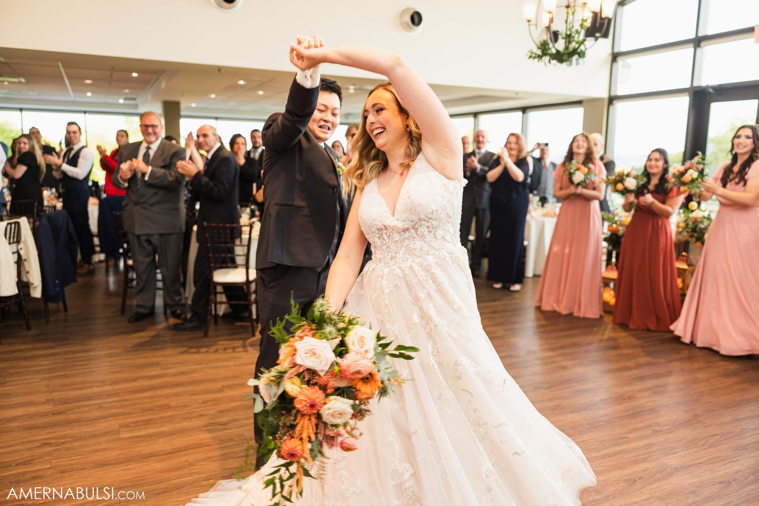 Bride in white gown and groom dance while guests celebrate at indoor wedding reception.