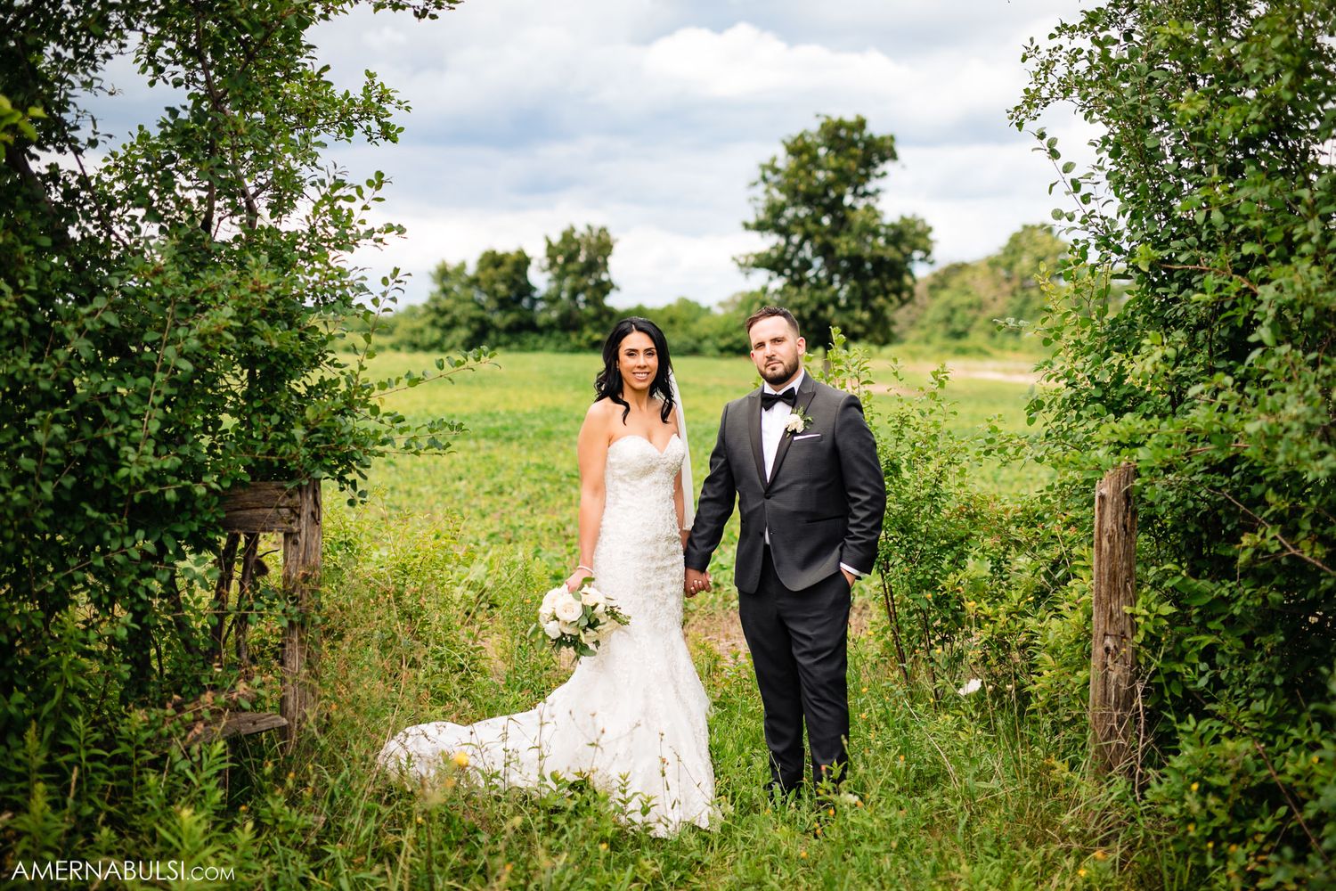 Elegant wedding couple poses in lush green meadow with flowing white dress and formal attire.