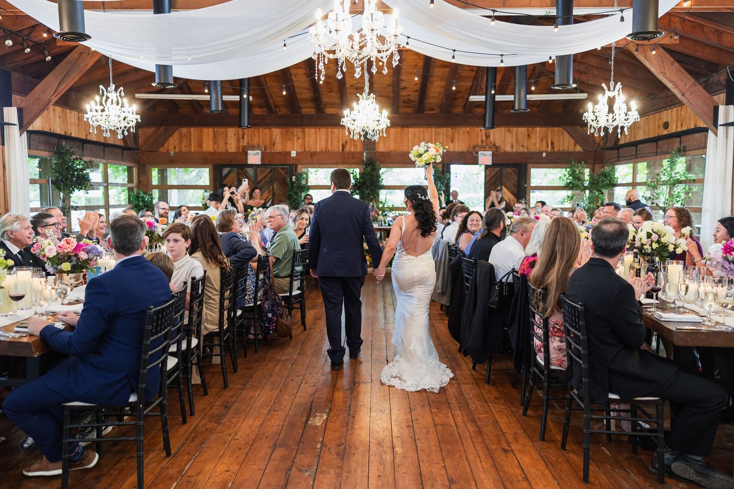 Bride and groom walking hand in hand into elegant rustic barn reception hall with chandeliers and seated guests.