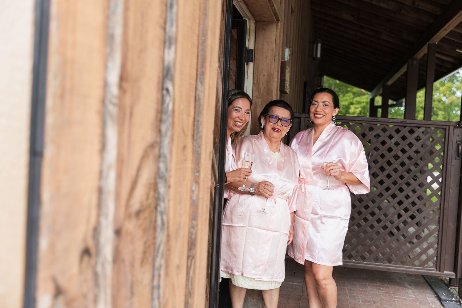 Three women in pink satin robes holding champagne glasses smiling in doorway.