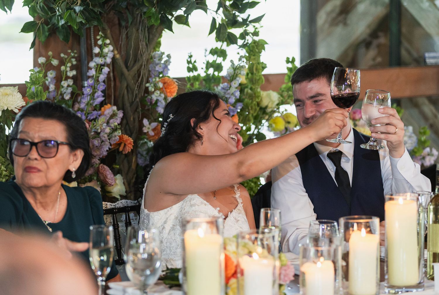 Bride and groom toast with red wine at wedding reception table with candles and flowers.