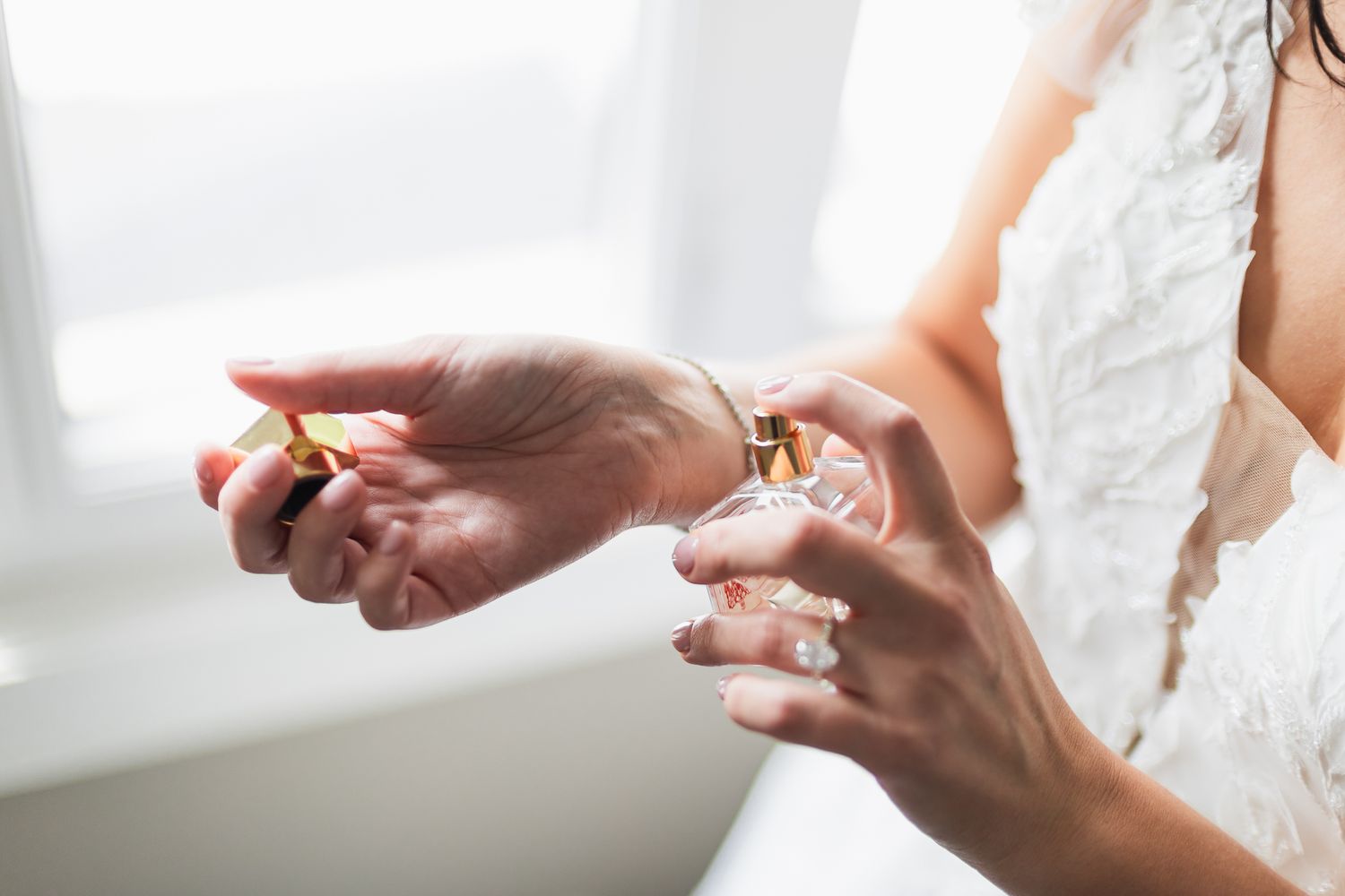 Bride applying perfume on her wrist while wearing a white lace wedding dress and gold bracelet.