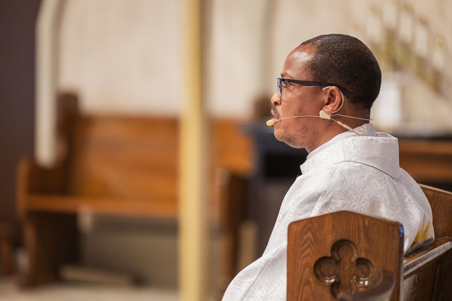 Catholic priest wearing white vestments and a microphone sits in contemplative prayer in a wooden church pew.