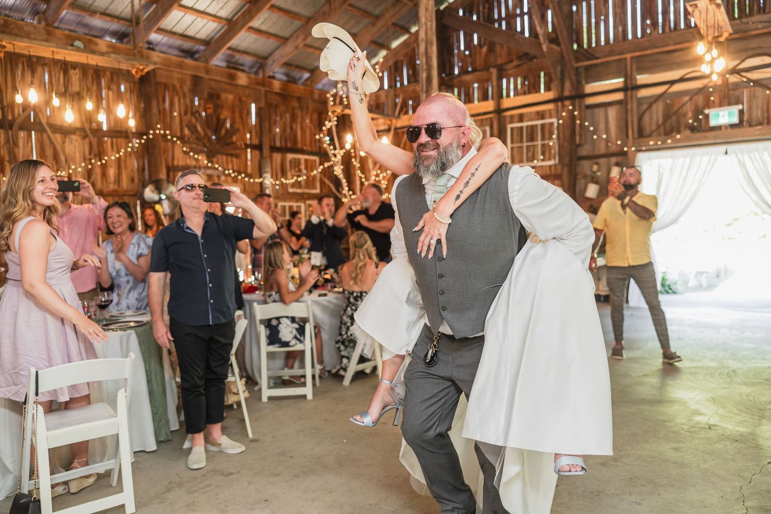 Groom carries bride into rustic barn wedding reception as guests cheer and celebrate.
