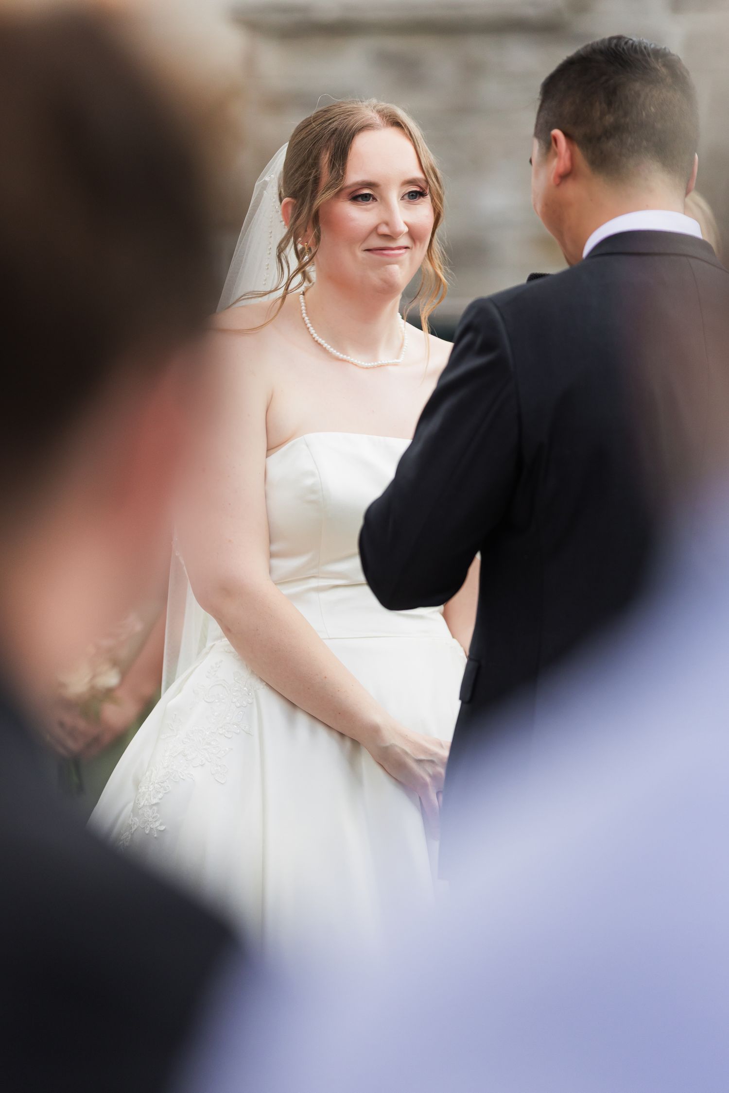 Emotional bride in strapless white gown and veil smiles at groom during wedding ceremony.