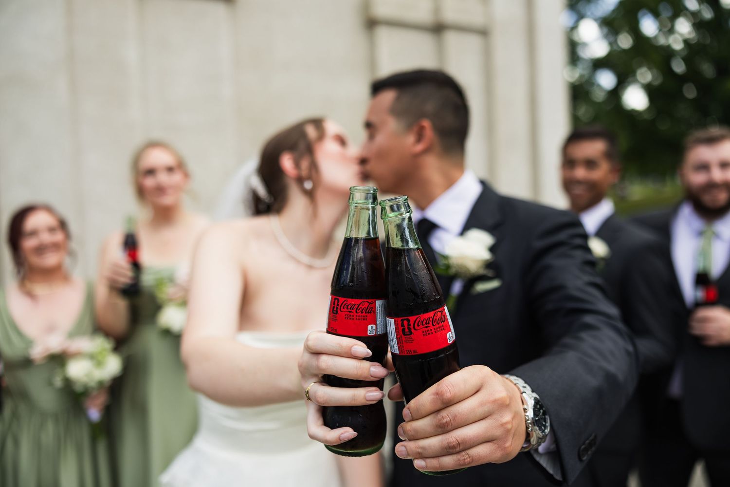 Wedding party toasting with Coca-Cola bottles as bride and groom share a kiss.