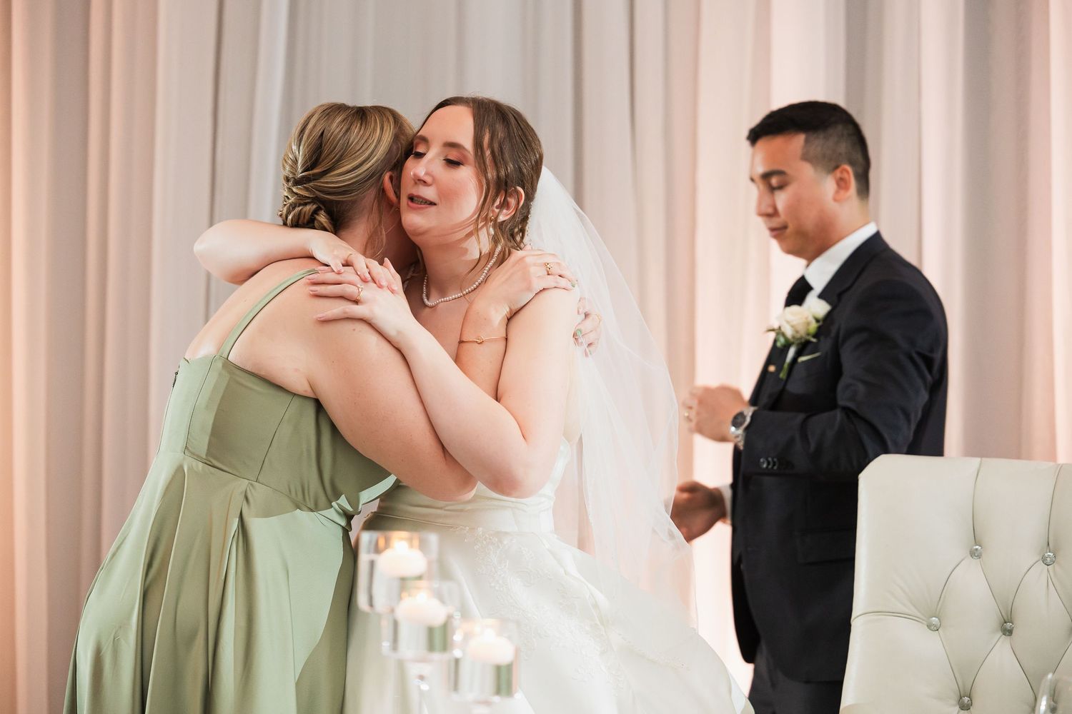 Bride hugging bridesmaid in green dress during wedding ceremony while groom stands nearby.