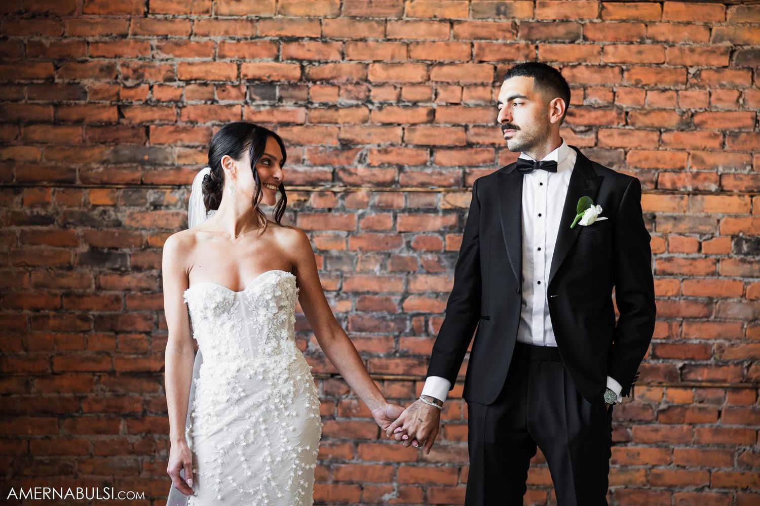 A couple poses against a rustic brick wall backdrop in formal wedding attire, holding hands.