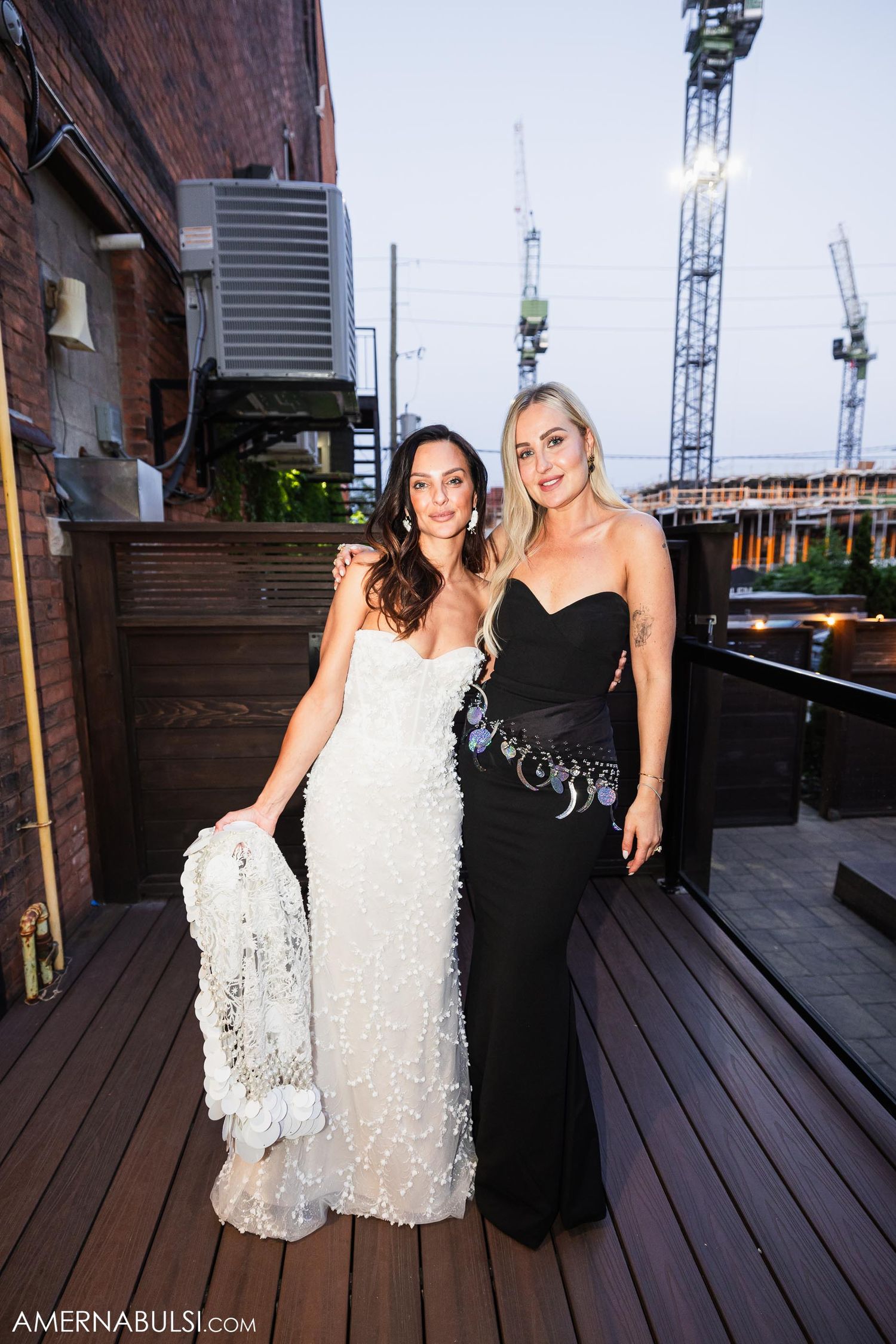 Two women in elegant dresses pose on outdoor deck with construction cranes in background.