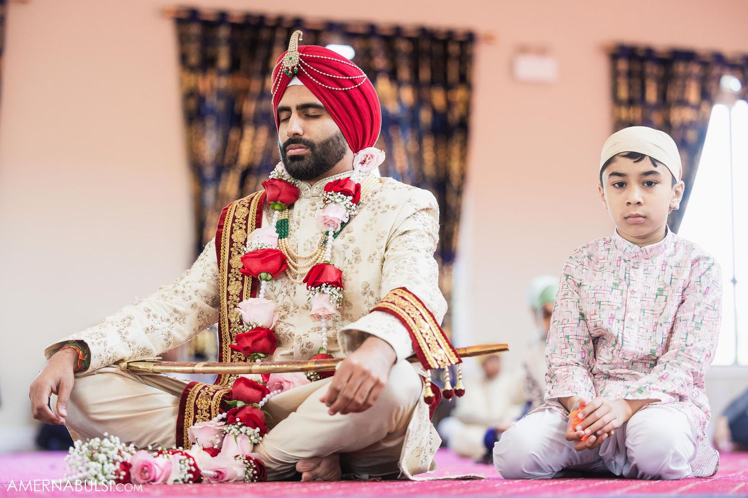 Traditional Sikh ceremony participants seated on pink carpet wearing formal attire and turbans.