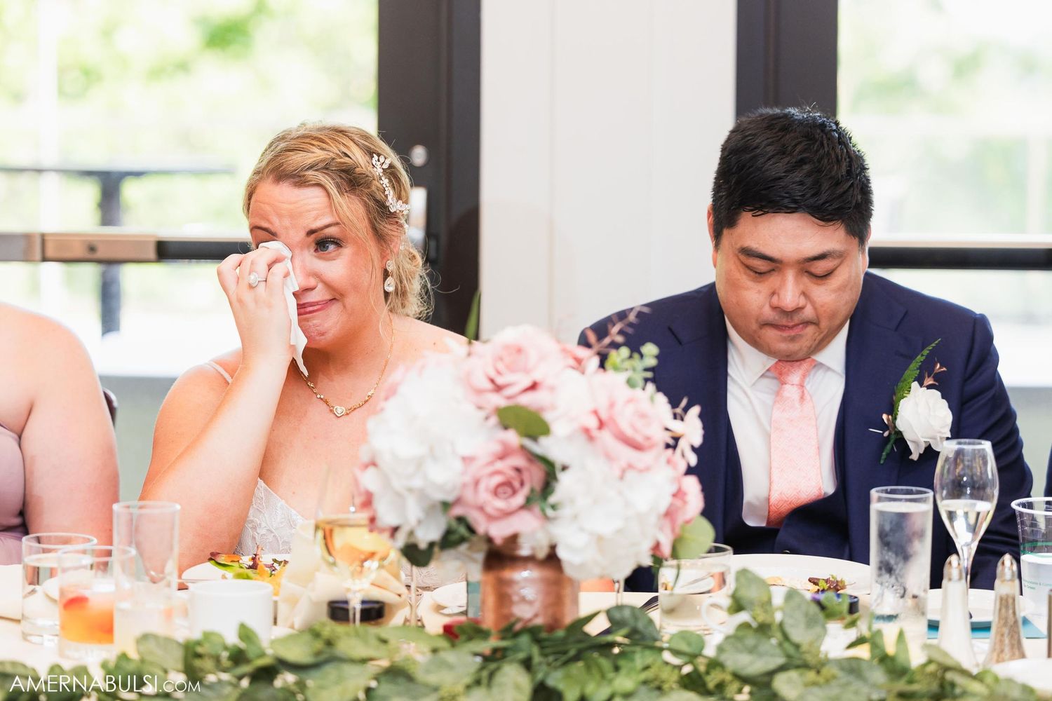 Wedding couple sits at decorated reception table with pink floral centerpieces and greenery garland.