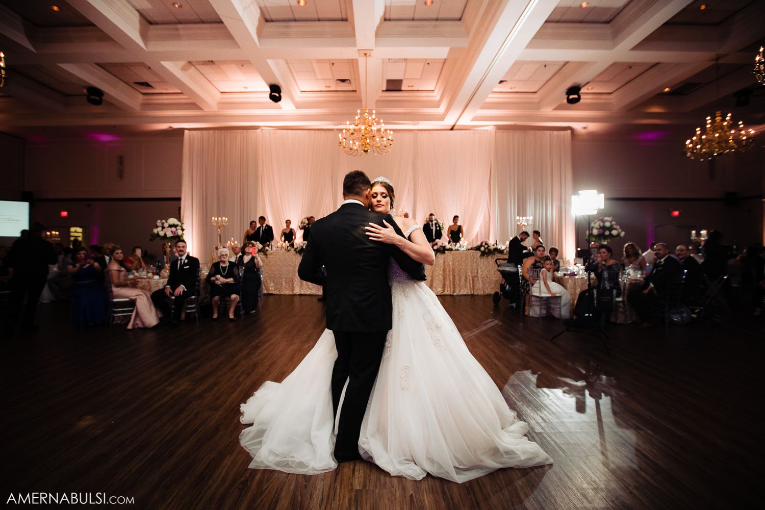 Wedding couple dances in spotlight on wooden dance floor in elegant ballroom setting.