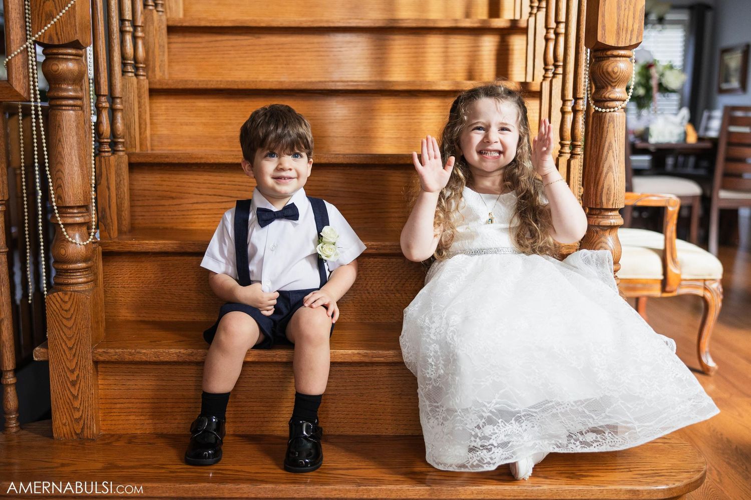 Two young children in formal attire sitting on wooden stairs, one in a bowtie and one in a white dress.