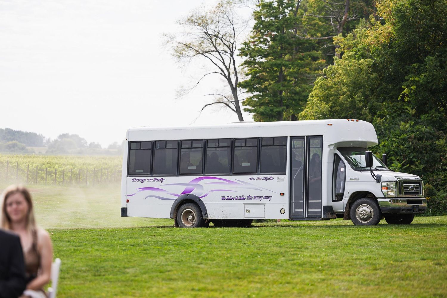 White shuttle bus parked on grassy field near trees at outdoor wedding venue.