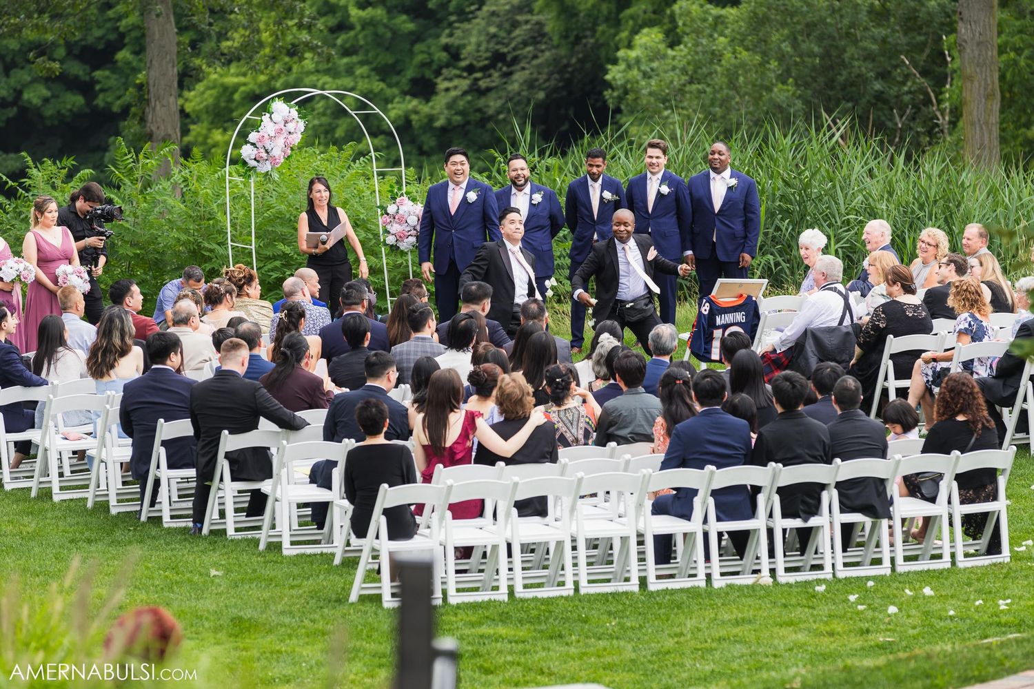 Wedding guests seated in white chairs on a lawn with wedding party standing under a decorative arch.