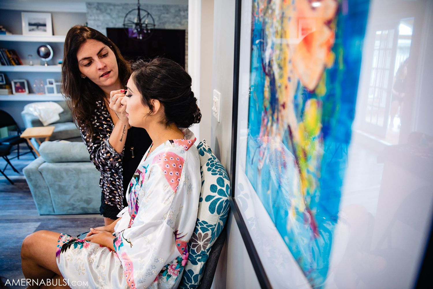 Two women in a room getting ready with makeup application while colorful artwork hangs on the wall.