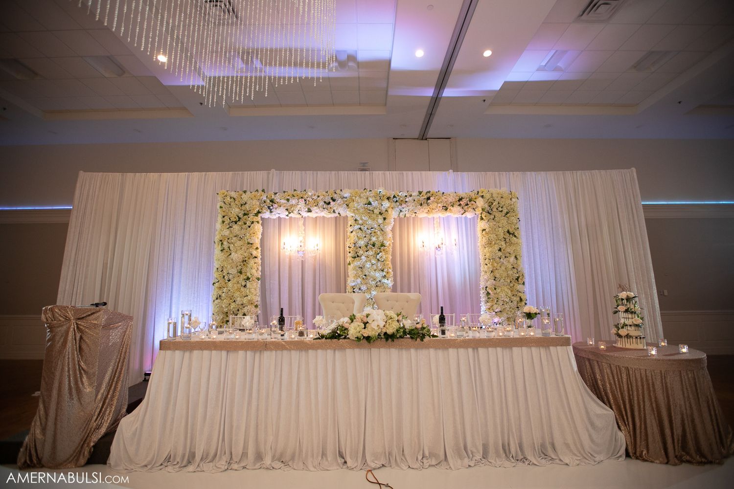 Luxurious white wedding reception decor with floral arrangements and draped fabric across head table.