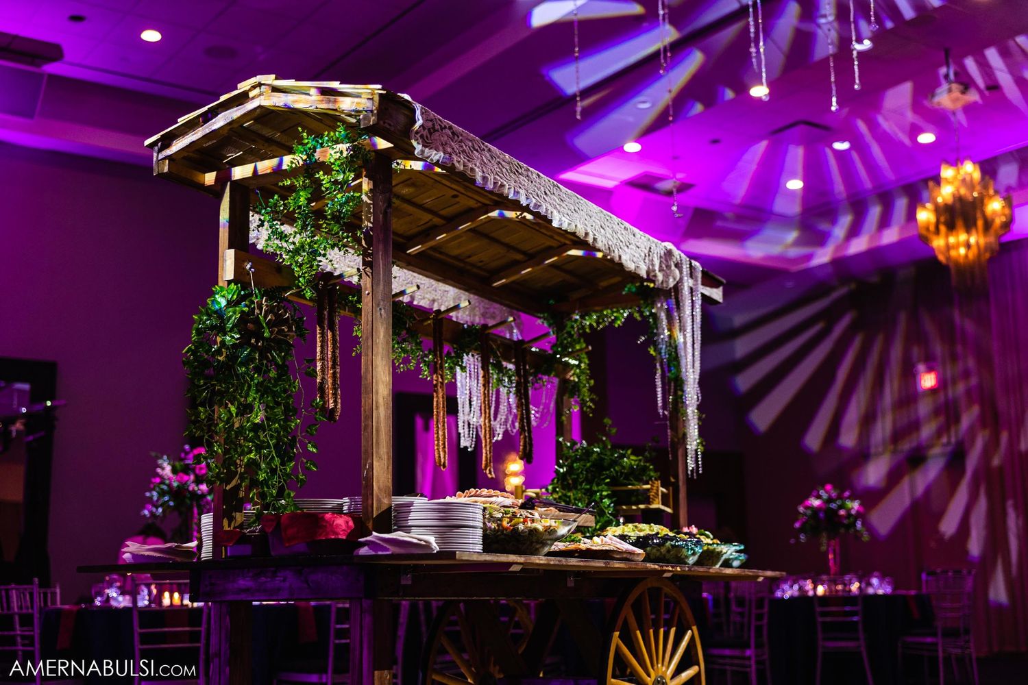 Wooden dessert cart decorated with greenery and lights under purple reception lighting.
