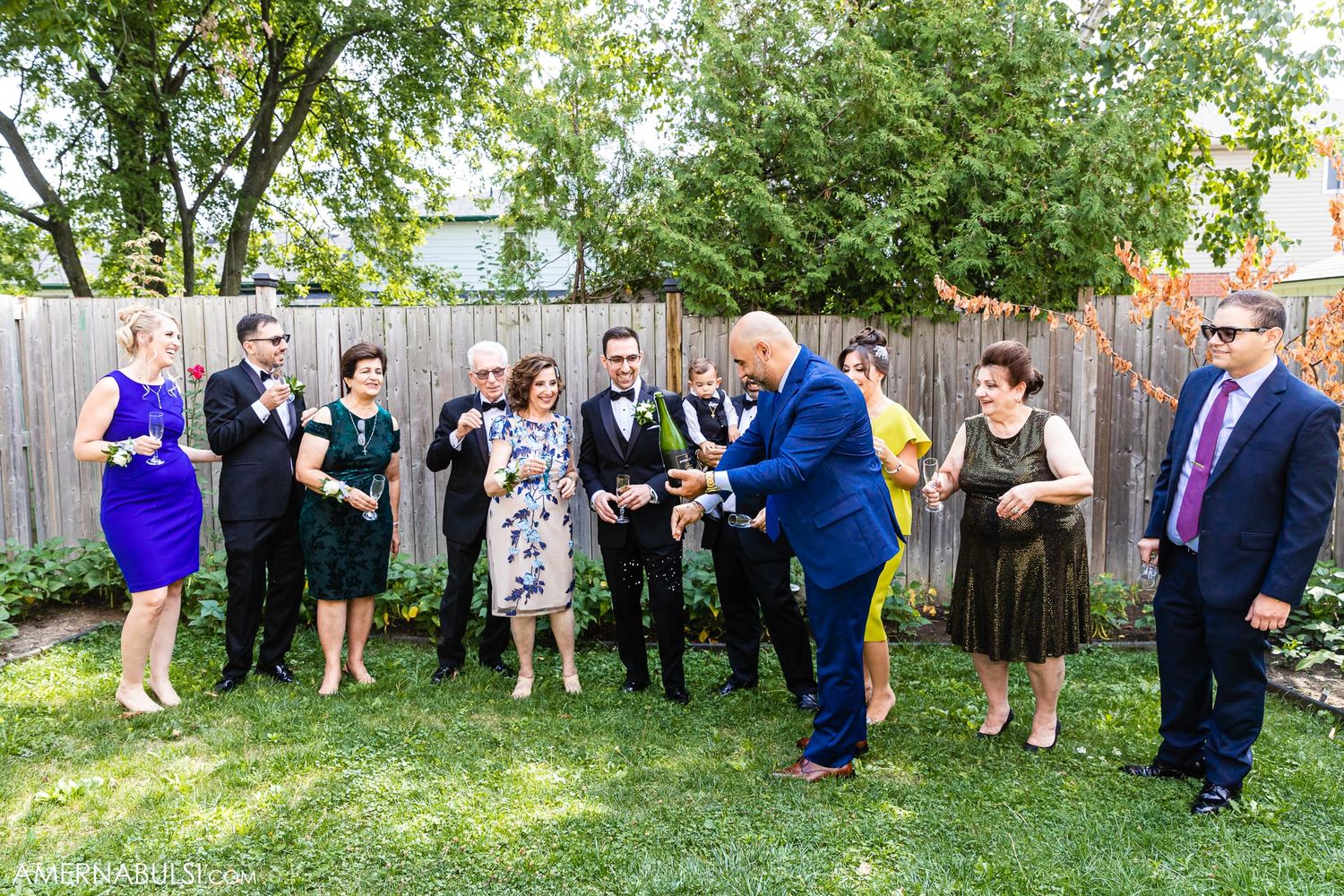 Group photo of wedding party in mixed formal attire standing in garden with wooden fence.