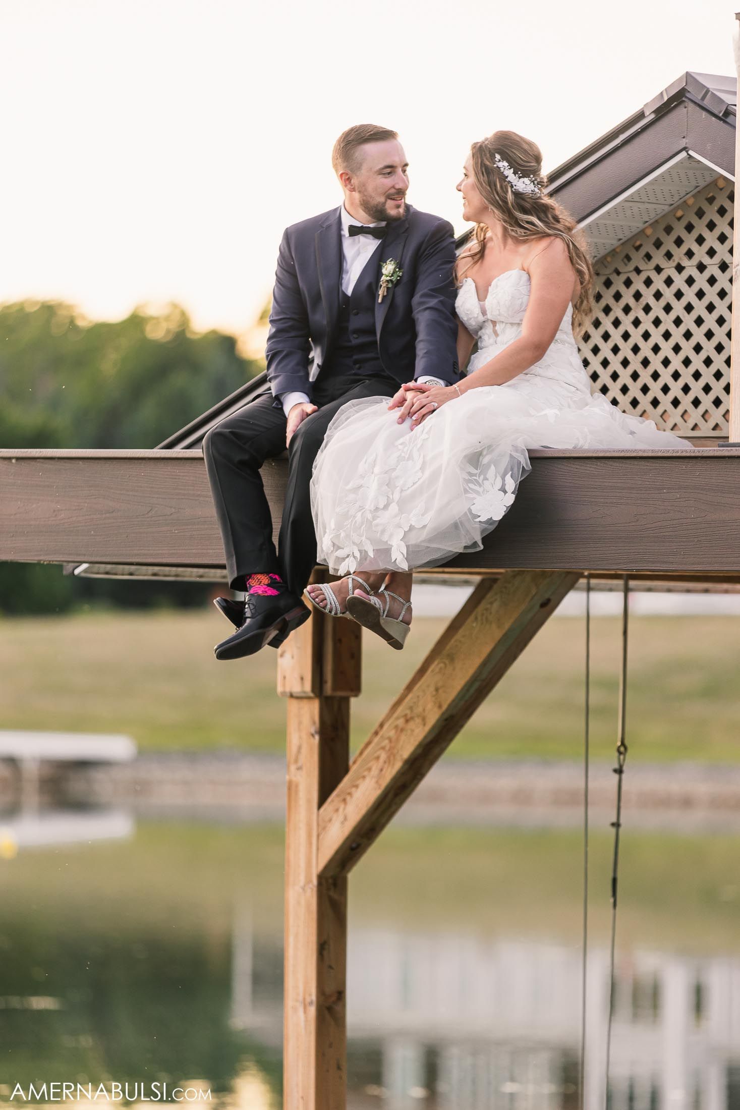 A romantic wedding couple sits together on a wooden deck railing at sunset near a lake.