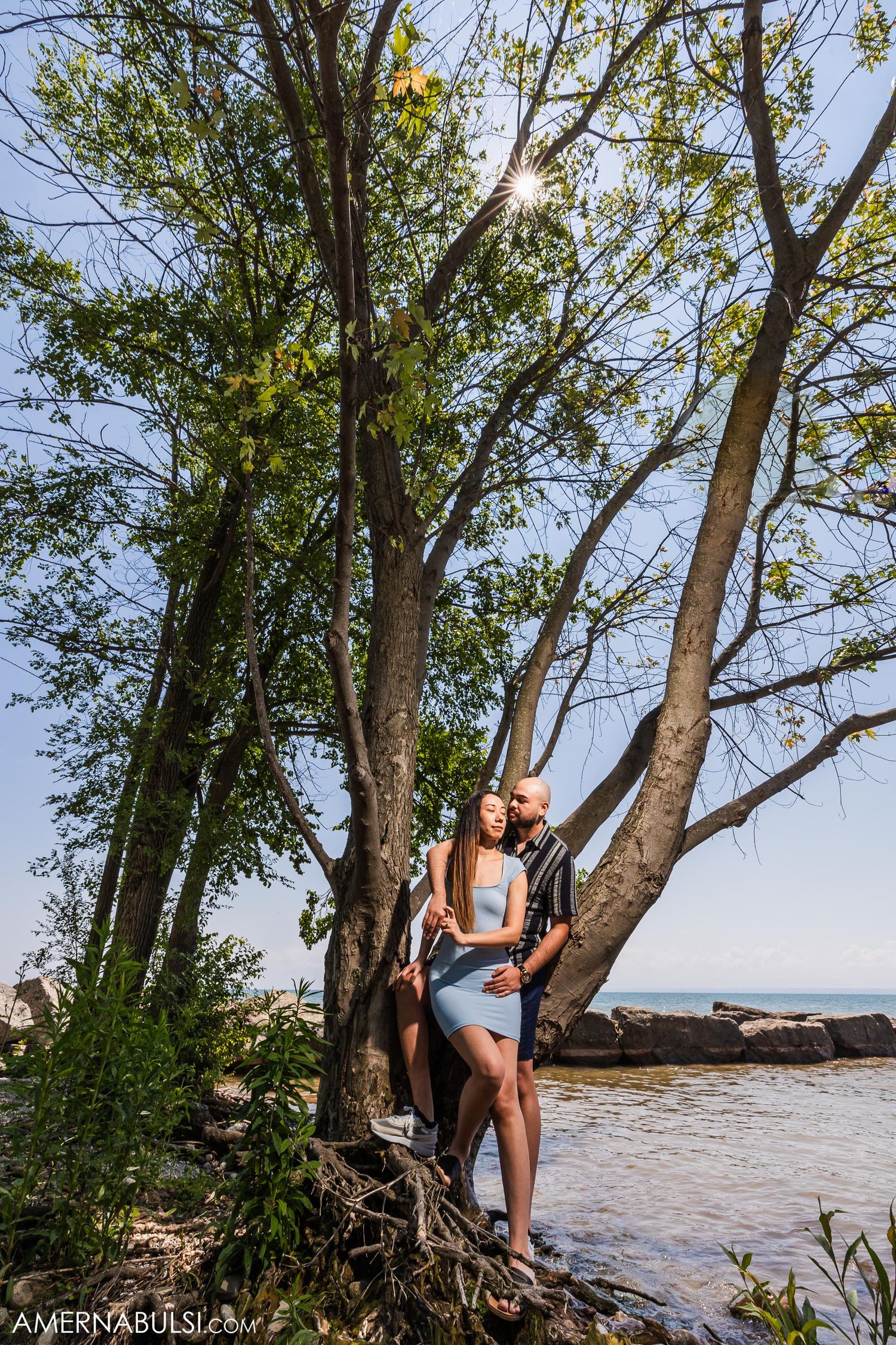 A couple stands among large tree branches near the water's edge.