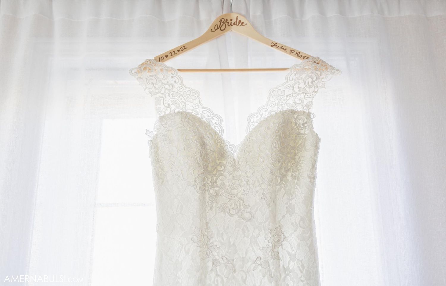 Delicate white lace wedding gown hanging against bright window backdrop.