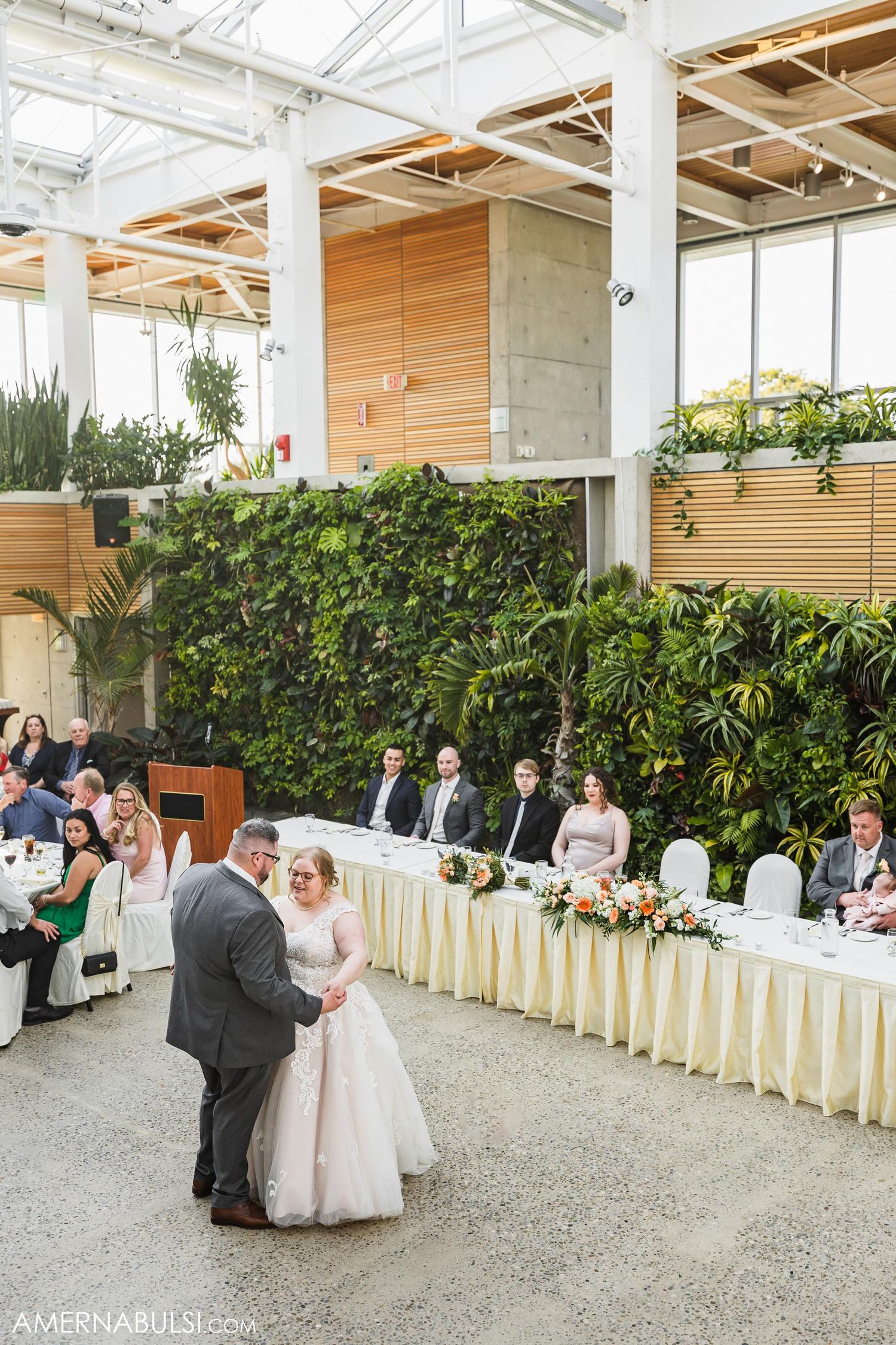 A couple shares their first dance at a modern indoor wedding venue with wooden walls and hanging plants.
