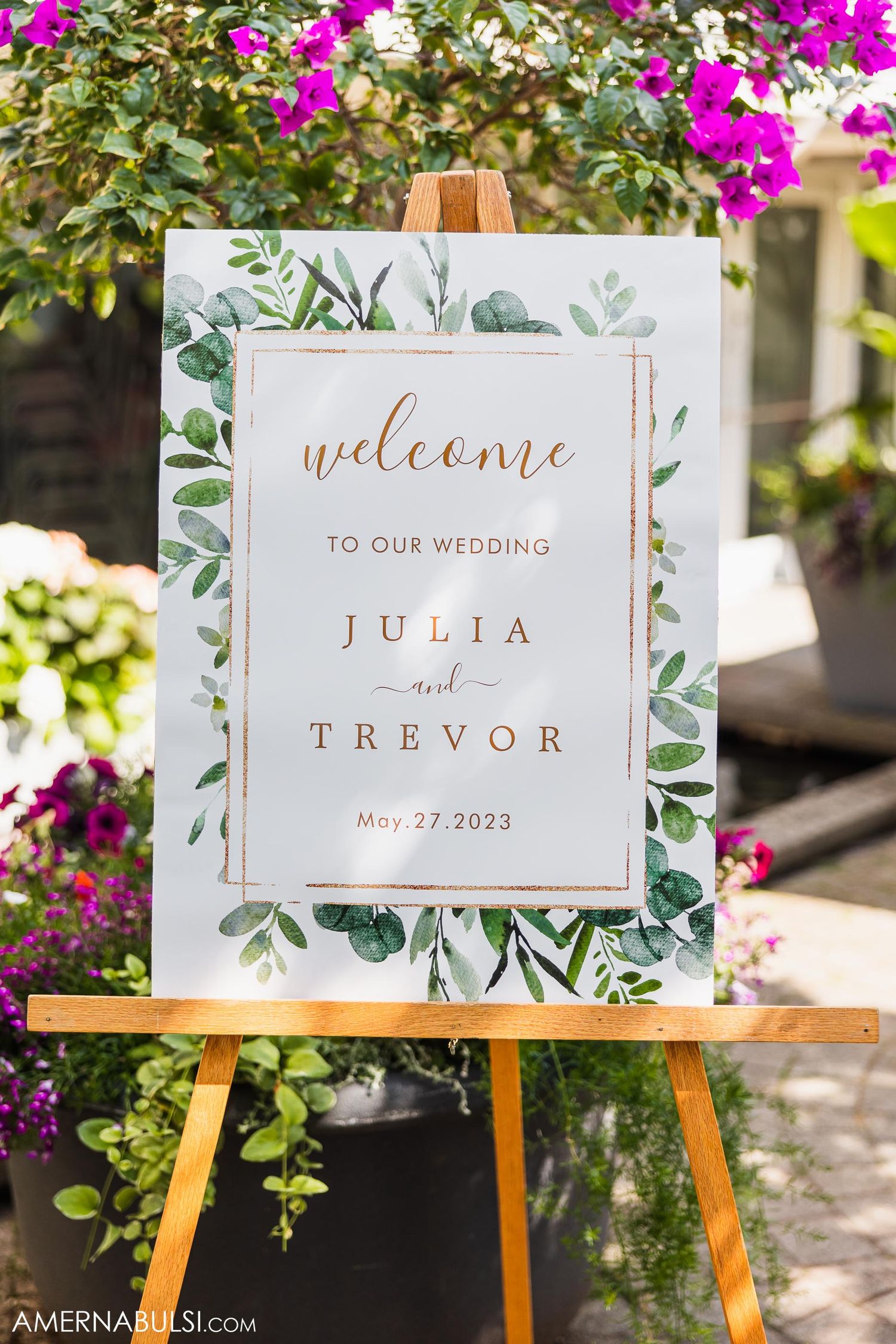 Elegant wedding welcome sign with greenery design on wooden easel surrounded by pink flowers.