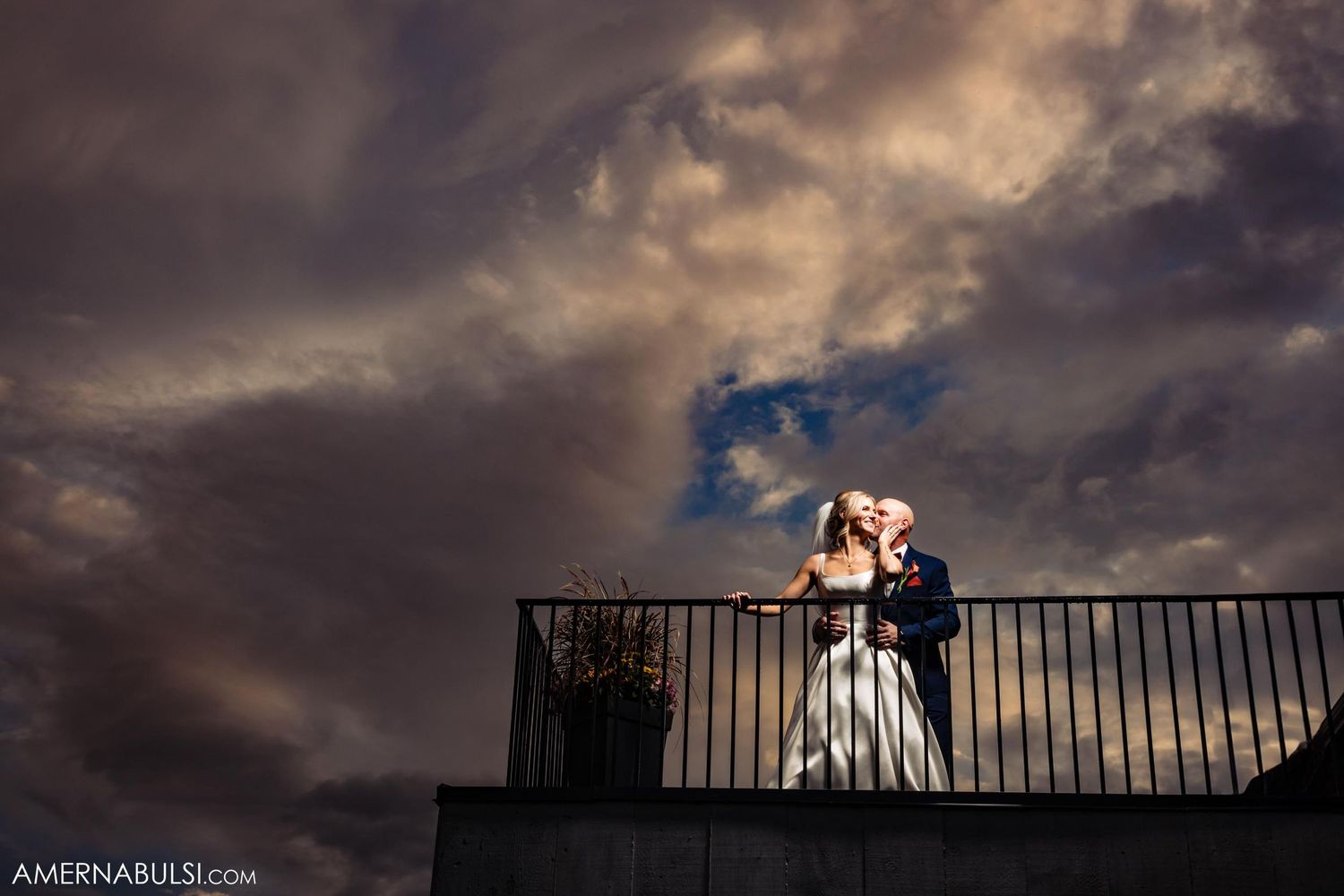 Silhouetted couple embraces on balcony against moody sunset clouds.