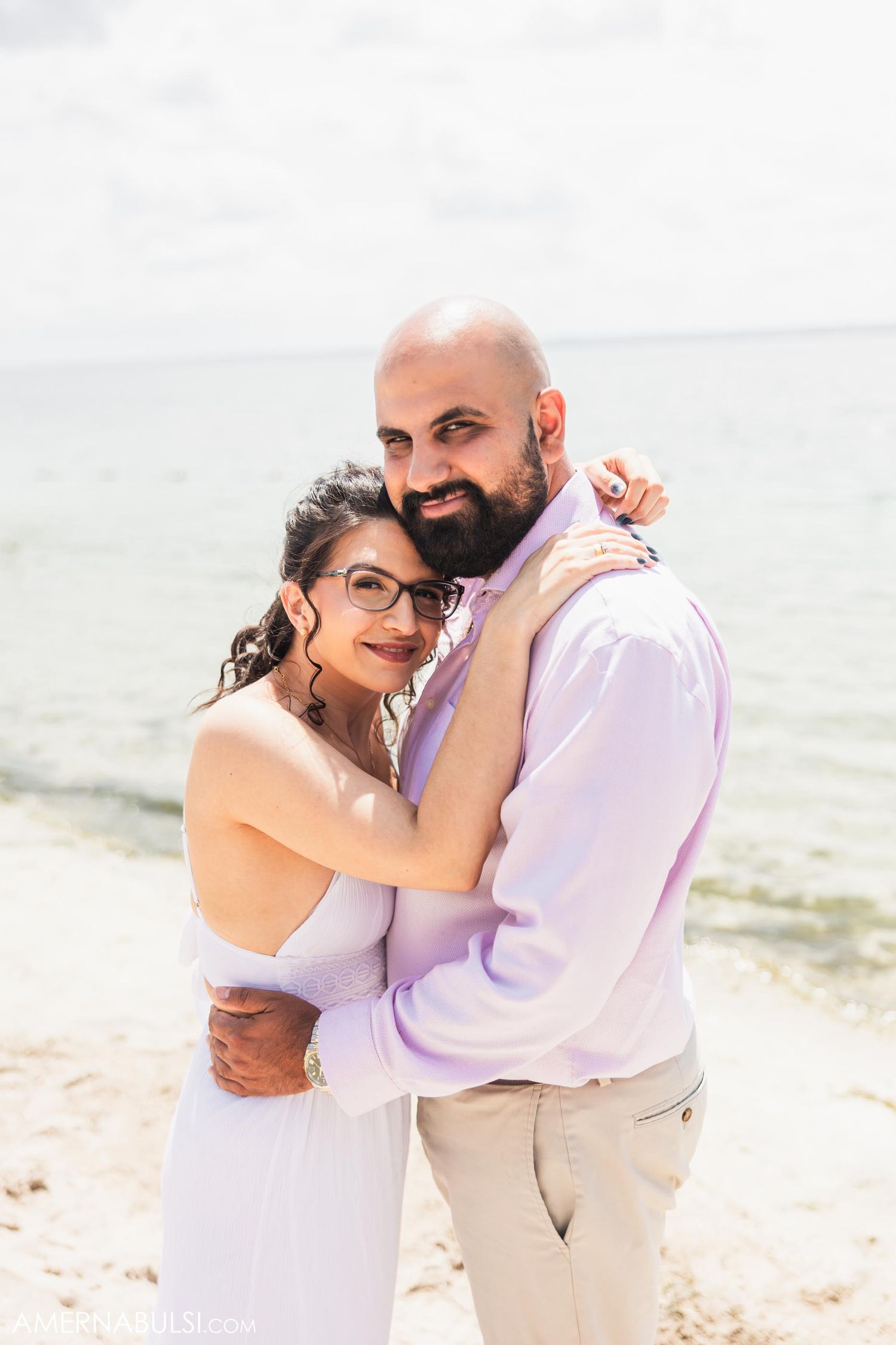 A couple embraces on a sunny beach in formal attire.