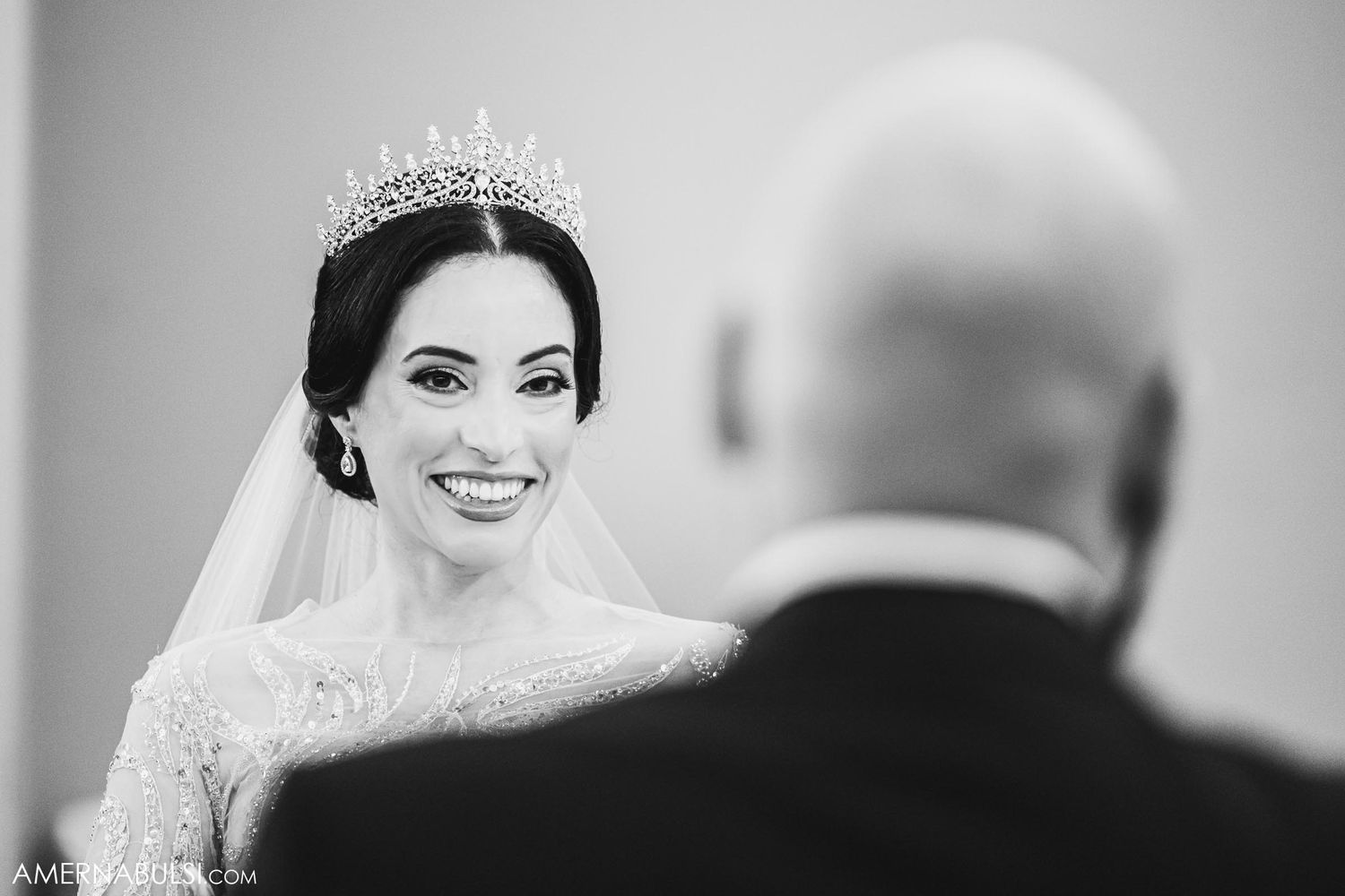 A bride wearing a sparkling tiara and white lace gown smiles during her wedding ceremony.