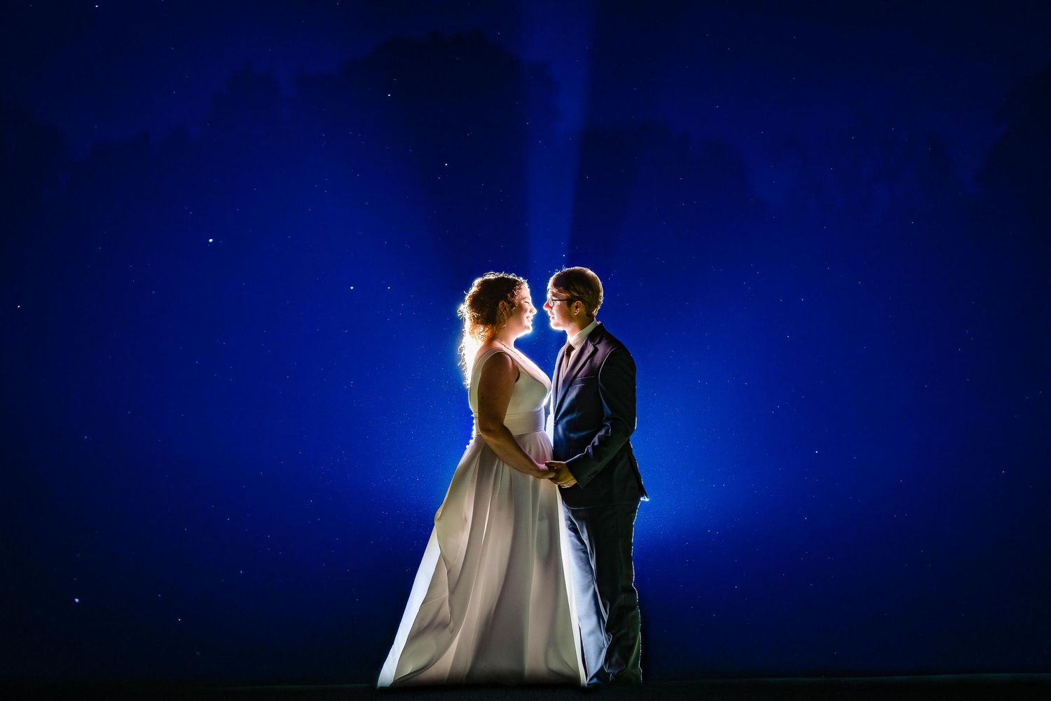 Silhouetted couple stands against dramatic deep blue night sky with backlit glow creating romantic atmosphere.