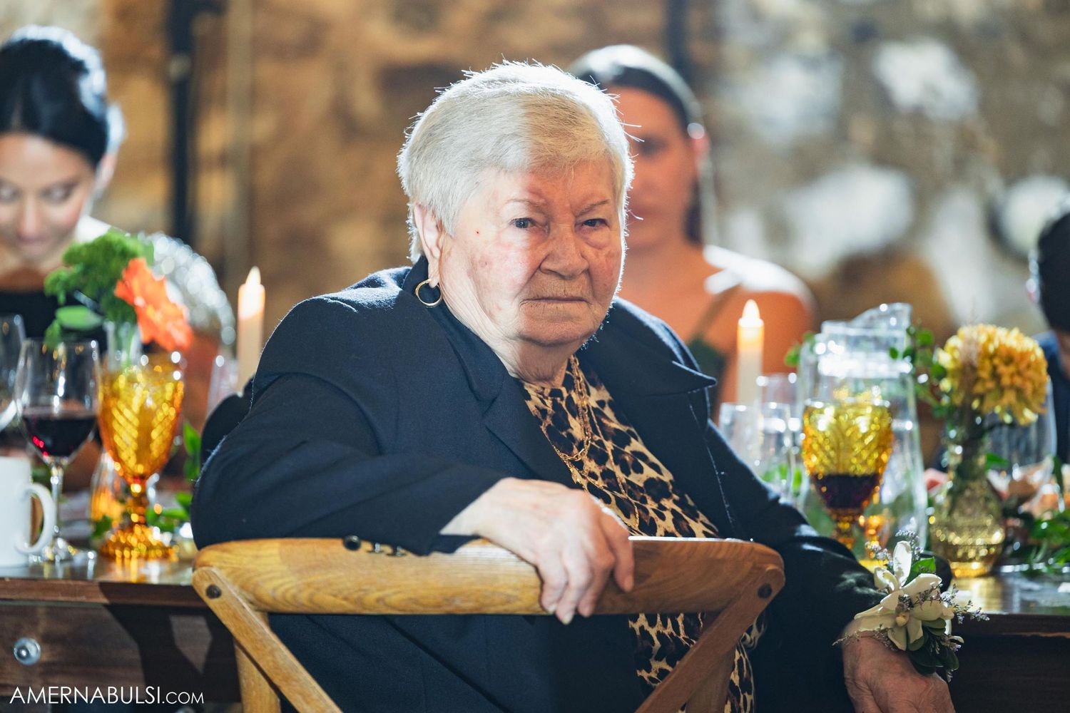 An elderly guest sits at a wedding reception table with drinks and floral centerpieces.